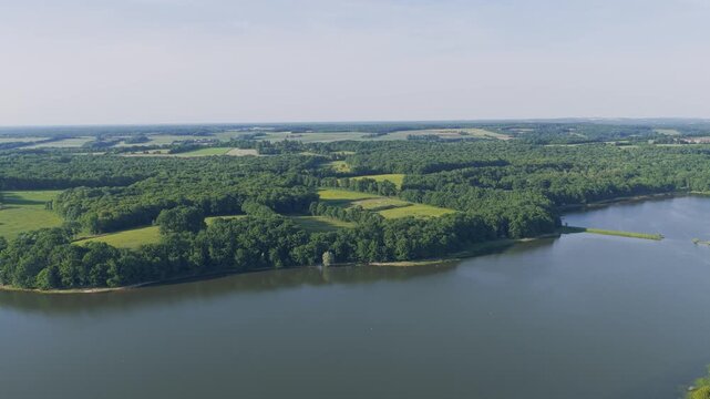 Aerial footage showcasing the expansive Lac du Bourdon surrounded by lush green forests and open fields in the Burgundy region of France. The video captures the tranquil lake, dense woodlands, and