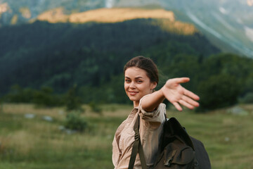 Obraz premium Young woman traveler with backpack smiles and reaches out hand inviting to join her in scenic mountain landscape with green meadows and forest background.