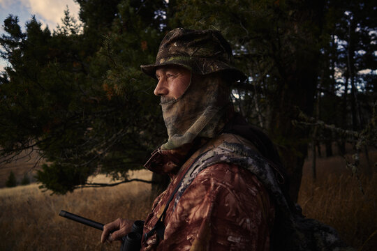 Hunter stands near trees with rifle at dusk in the forest
