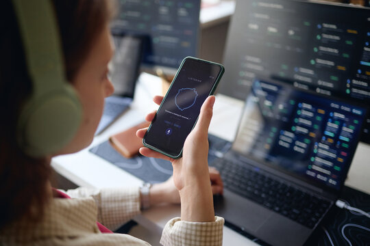 Young adult woman holding smartphone and using voice assistant while working at laptop with data dashboards, showing multitasking and AI productivity. Ideal for office work or tech marketing