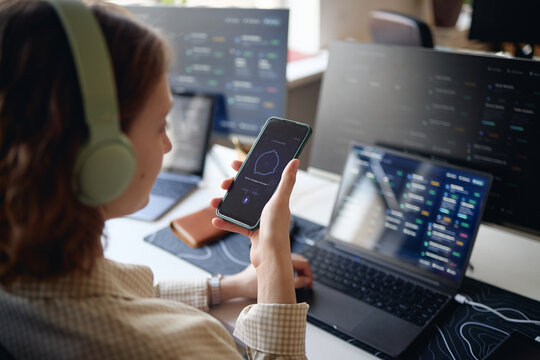 Young adult woman holding smartphone and monitoring digital dashboards across laptops while wearing headphones, managing workflow and multitasking. Ideal for office work apps or productivity marketing