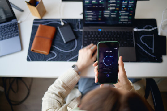 Overhead shot of young adult woman using smartphone voice assistant while working on laptop at desk, multitasking across devices. Ideal for AI productivity, digital workflow, smart tech marketing