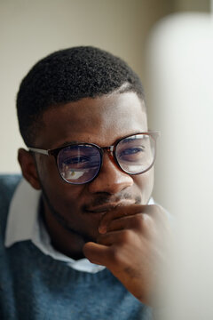 Vertical shot of young Black man wearing glasses, focusing on computer screen and resting chin on hand, suggesting analysis and planning. Ideal for business strategy or tech work