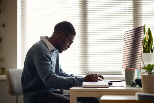 Medium side view of young adult Black man working at desk, writing notes beside desktop monitor, focusing on planning and productivity. Ideal for office work, business