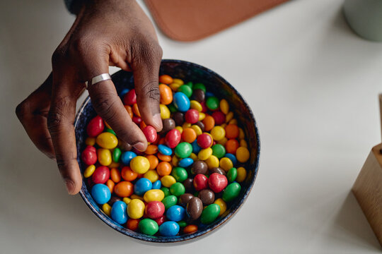 Close up of Black adult hand picking colorful chocolate candies from bowl, suggesting snacking and indulgence. Ideal for confectionery ads, sugar consumption, retail promotion