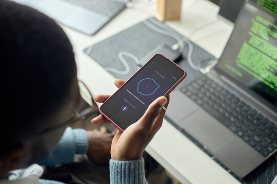 High angle of man holding smartphone and using voice assistant beside laptop with code, showing digital productivity. Ideal for tech, app, automation marketing