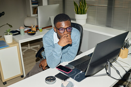 High angle shot of young Black man working at computer, thinking through task in office workspace. Ideal for business productivity, focus, remote work, burnout themes