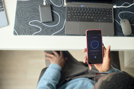 Overhead shot of Black man holding smartphone and using voice assistant beside laptop at desk, demonstrating mobile productivity. Ideal for AI app, remote work, digital service marketing