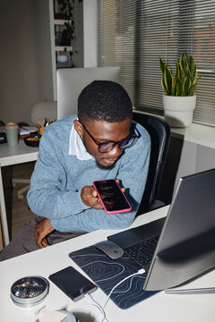 Vertical shot of young adult Black man holding smartphone near mouth while working at laptop, recording voice message or joining call. Ideal for remote work, mobile communication, productivity
