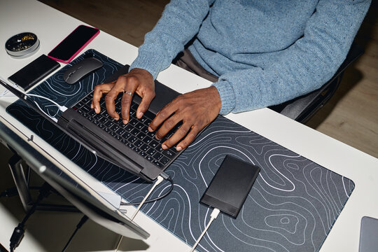 Black man typing on laptop at desk, charging devices and managing digital workflow in office. Ideal for office work, productivity, business, top view