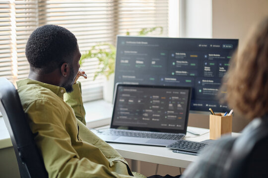 African American man reviewing analytics dashboard on desktop and laptop during team discussion. Ideal for business intelligence, fintech, workflow software marketing