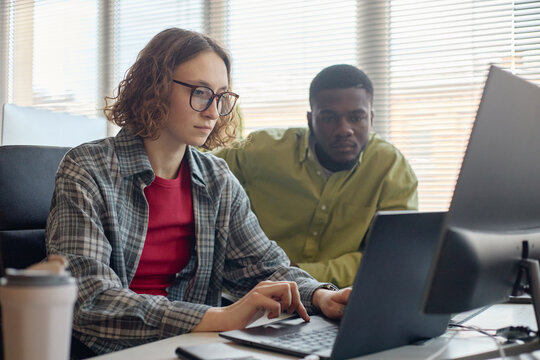 Young adult Caucasian woman working on laptop while Black man reviewing screen in office, collaborating on project and problem solving. Ideal for teamwork, mentoring, workplace training