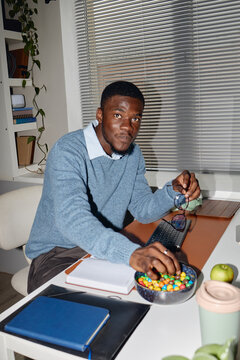 Portrait of young Black man working at desk, reaching for candy while holding eyeglasses near laptop and notebook. Supporting work, productivity break, office lifestyle marketing