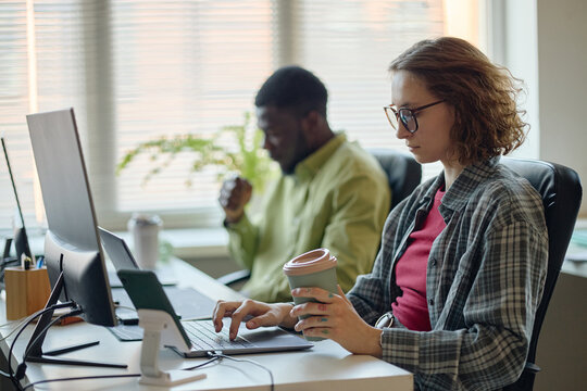 Young adult Caucasian woman working on laptop while holding coffee in office, Black man collaborating in background. Useful for workplace productivity, startup team, hybrid work marketing