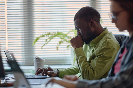 Medium shot of African American man working on laptop, reviewing data with focused colleague in office. Ideal for teamwork, productivity, corporate planning campaigns