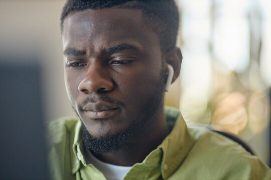 Close up of young Black man wearing wireless earbuds, focusing on computer screen while working online, conveying concentration. Ideal for remote work, productivity, tech use