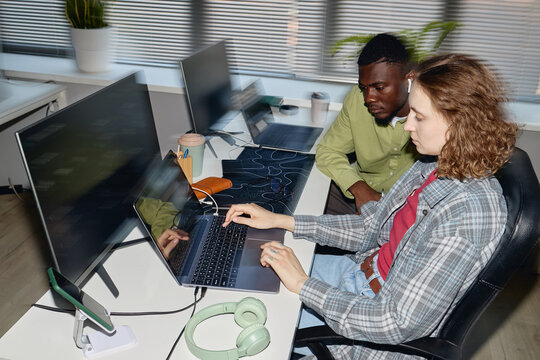Young adult coworkers collaborating at laptop workstation, reviewing digital project and discussing workflow in office. Ideal for teamwork, startup, productivity, tech business content