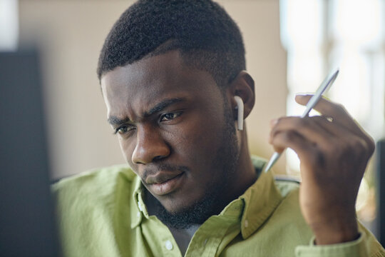 Close up of young Black man wearing wireless earbuds, concentrating on computer work and holding stylus. Ideal for remote work, online learning, productivity or focus concept