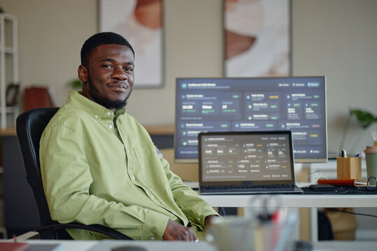 Portrait of young Black man sitting at workstation, monitoring business analytics on laptop and desktop screens. Ideal for data analysis, remote work, medium shot