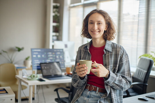 Portrait of young adult Caucasian woman holding coffee mug and smiling in office, representing workplace confidence and creative routine. Ideal for remote work or startup marketing