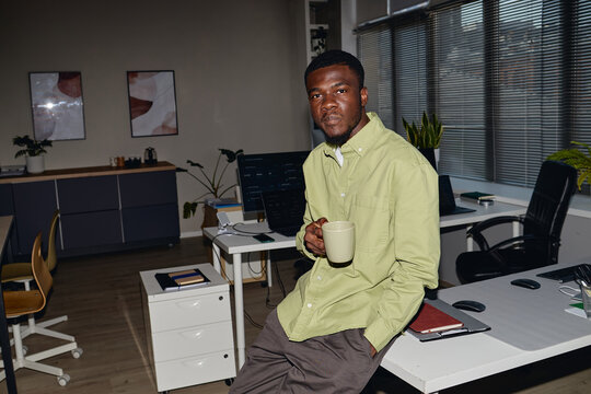 Portrait of young Black man holding coffee mug while sitting on office desk, taking work break in modern workplace. Ideal for business productivity, office culture, employee wellbeing