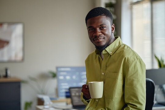 Portrait of young Black man holding coffee mug, looking at camera in office workspace, conveying confidence and professionalism. Ideal for career growth, workplace branding, team culture