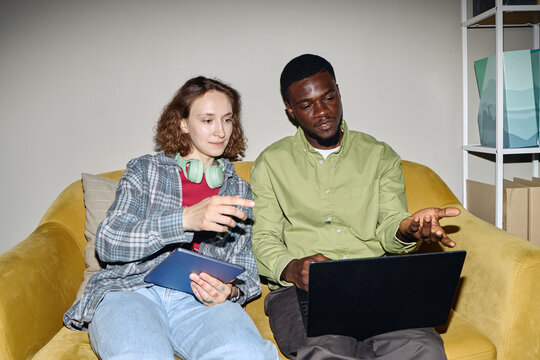 Young Caucasian woman and Black man collaborating on laptop and tablet while sitting on sofa, discussing project workflow and teamwork. Useful for remote work, startup, training marketing