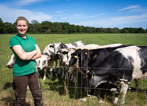 Eine junge Landwirtin in braun-gr&uuml;ner Arbeitskleidung steht l&auml;chelnd in Ganzk&ouml;rperansicht auf einer gr&uuml;nen Sommerweide, w&auml;hrend direkt hinter ihr rot-bunte Holstein-K&uuml;he grasen.