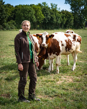 A young female farmer in workwear stands smiling on a green summer meadow in a full-length shot, with red-and-white Holstein dairy cows grazing directly behind her.