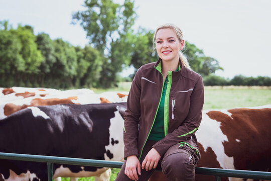 Eine l&auml;chelnde junge Landwirtin in Arbeitskleidung sitzt auf einem gr&uuml;nen Metall-Weidetor, w&auml;hrend im Hintergrund eine Gruppe von schwarz-bunten und rot-bunten Milchk&uuml;hen auf der Koppel steht.