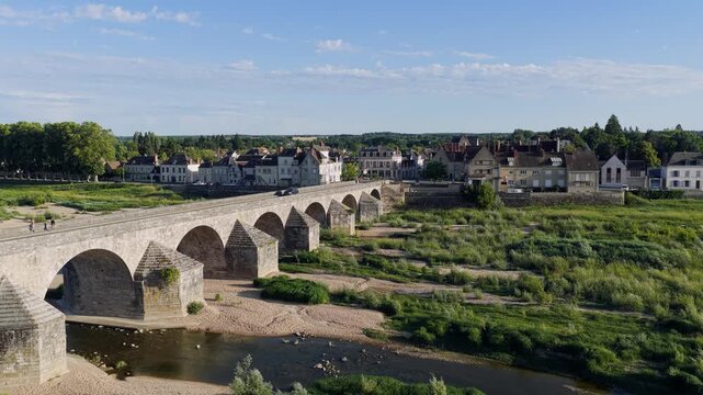 Drone footage capturing a panoramic view of the historic stone bridge spanning the Loire River in Gien, France. The scene shows the bridge s arches, the riverbanks with green vegetation, and the