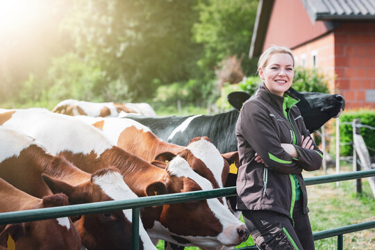 Eine l&auml;chelnde junge Landwirtin in Arbeitskleidung sitzt auf einem gr&uuml;nen Metall-Weidetor, w&auml;hrend im Hintergrund eine Gruppe von schwarz-bunten und rot-bunten Milchk&uuml;hen auf der Koppel steht.