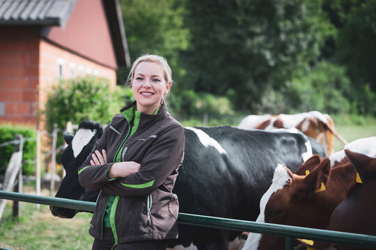 Eine junge Landwirtin in braun-gr&uuml;ner Arbeitskleidung steht mit verschr&auml;nkten Armen an einem gr&uuml;nen Metallzaun, w&auml;hrend im Hintergrund ihre Herde aus schwarz-bunten und rot-bunten K&uuml;hen steht.