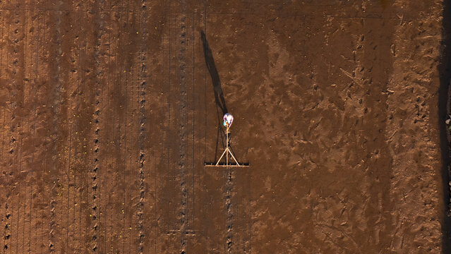 Aerial view of a farmer working in a muddy field with a wooden rake tool, casting a long shadow on the soil in Wukirsari, Daerah Istimewa Yogyakarta, Indonesia.