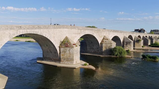 A wide daytime view of a historic stone arch bridge spanning the Loire River in Gien, France. The video shows the bridge s multiple arches, calm river water below, and a clear blue sky. Occasional
