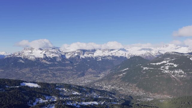 Wide panoramic aerial view of the snow-covered Alps mountains and forested hills overlooking the Sallanches valley in the French Alps. The scene features clear blue skies, scattered clouds, and