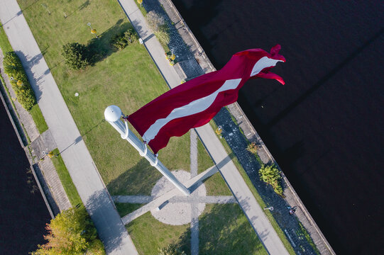 Aerial view of the massive Latvian flag flying on the AB Dambis flagpole, surrounded by green park paths and the Daugava river in Riga, Latvia.
