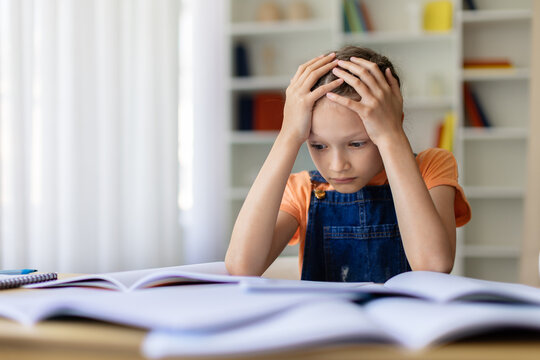 Teen girl holding her head while studying with open books at home, showing homework stress, academic pressure and frustration during school assignments