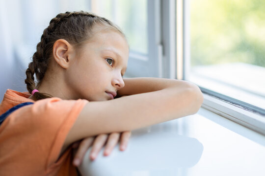 Pensive teen girl leaning on windowsill at home and looking outside, showing loneliness, boredom or quiet reflection in a close emotional lifestyle portrait
