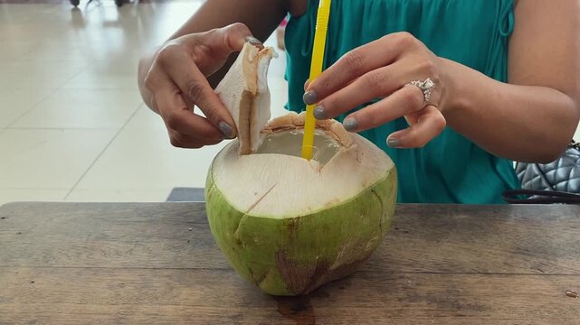 Asian Woman Drinking Fresh Young Coconut Water Juice with Straw, Sitting and Relaxing in Open Street Food Market with Face Mask Pulled Down