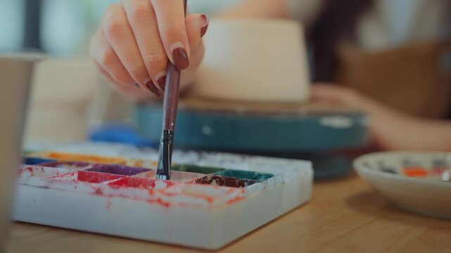Close up of young Asian woman hand holding paintbrush and dipping color palette to paint ceramic cup, creative female artisan decorating pottery in workshop studio, art class crafting hobby.