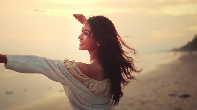 Happy Asian woman holding and playing with colorful smoke bombs on the beach during sunset. Young female enjoying summer vacation with blue and orange smoke flares. Creative celebration and freedom