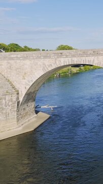 Daytime video of a historic stone arch bridge crossing a wide river in Gien, France. The camera moves along the bridge, showing the flowing water, green riverbanks, and clear blue sky. No people are