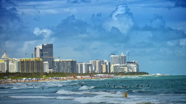 Crowded shoreline with swimmers and South Beach skyline, Miami Beach, Florida. Distant view of beachgoers, waves and highrise buildings under dramatic cloudy sky.