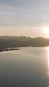 Aerial footage of sunrise over Lake Aiguebelette in Savoie, France. The scene captures the calm water, distant hills, and soft morning light reflecting on the lake surface. No people are visible