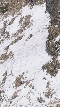 A wild chamois moves across a steep, snow-covered alpine slope at Col des Aravis in the French Alps. The rugged mountain terrain is partially covered with snow and exposed rock, highlighting the