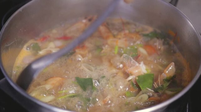 Spoon stirs Thai Tom Yum Goong hot & sour soup, simmering shrimp prawns & aromatic lemongrass, kaffir lime leaves herbs vegetables in boiling cooking pot. Asian home cooked meal