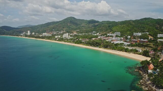 Amazing Karon Beach in Phuket, Thailand, long white sand shore, clear turquoise Andaman Sea, lush green hills, and beachfront hotels under a cloudy sky. Summer vacation travel destination. Aerial shot