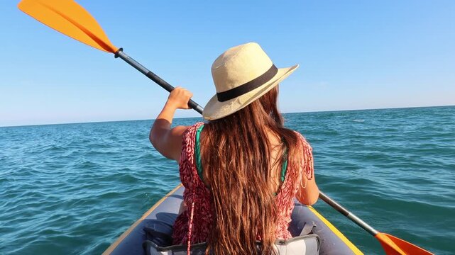 Active young woman with long hair and a hat paddling a kayak on the calm blue sea, enjoying a sunny day outdoors. Her back is to the camera as she rows through the water, embracing the scenery