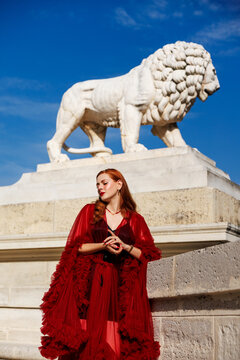 Fashion influencer Greta Gardner (Ivana Benko) posing in a dramatic red tulle dress next to a majestic stone lion statue at Place de la Concorde, Paris, under a bright blue sky.